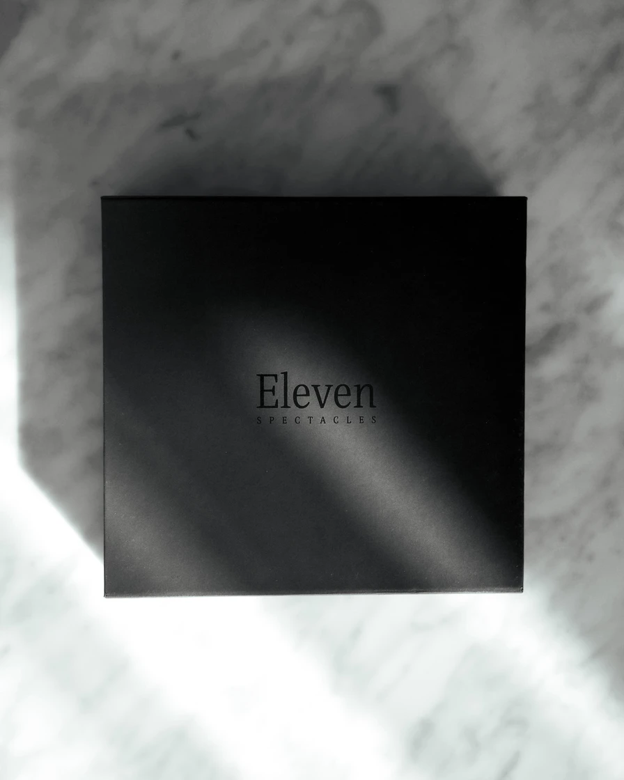 Black Eleven Spectacles eyewear box with a debossed logo, resting on white marble in soft diagonal light